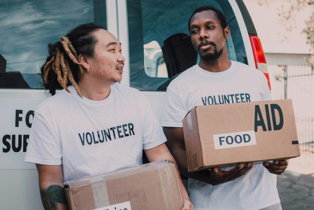 Two volunteers wearing white shirts distribute food aid boxes outdoors as part of a charity effort.